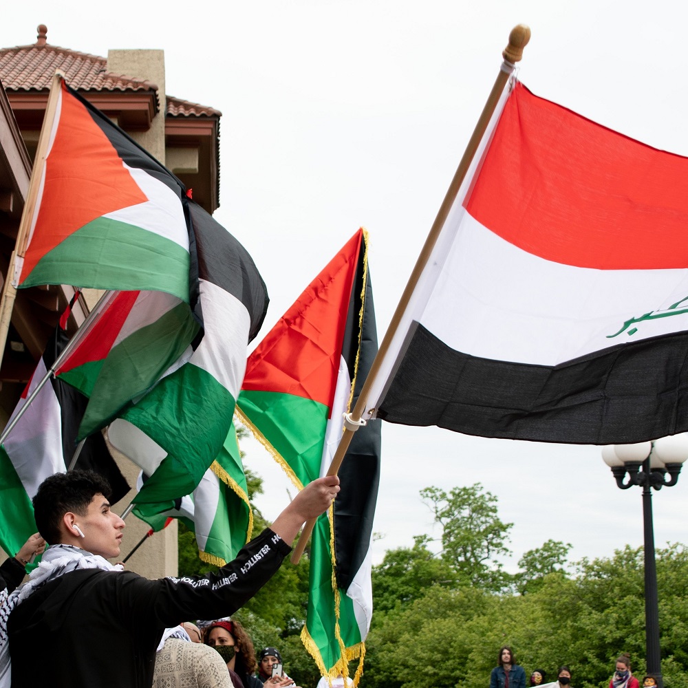 A protestor holding the flag of Iraq stands in solidarity at a protest demanding an end to Israeli apartheid and ethnic cleansing in occupied Palestine